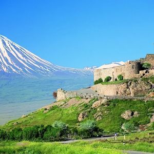 Eine steinerne Festung liegt auf einem grasbewachsenen, felsigen Hügel, mit einem großen, schneebedeckten Berg im Hintergrund vor einem klaren blauen Himmel.