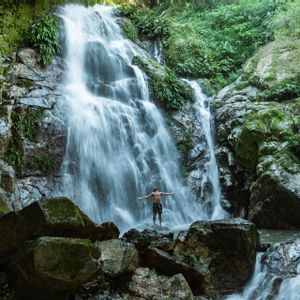 Ein Mann, oberkörperfrei und mit ausgestreckten Armen, sitzt auf den Felsen am Fuße eines Wasserfalls im Herzen eines dichten Dschungels.