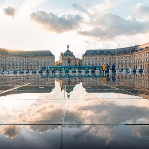 Un grande edificio classico e un cielo nuvoloso si riflettono nel terreno bagnato di una grande piazza pubblica al tramonto.