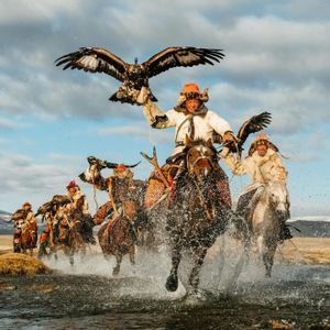 Cacciatori di aquile, in abiti tradizionali, galoppano a cavallo attraverso un fiume, con le aquile al braccio.