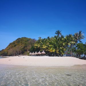 Vista dall'acqua limpida e poco profonda di un'isola tropicale con spiaggia di sabbia bianca, palme e una collina boscosa sotto un cielo blu.