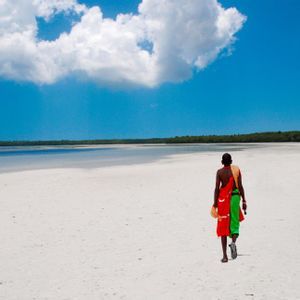 Una persona, vista di spalle, che indossa abiti tradizionali colorati cammina su una vasta spiaggia di sabbia bianca sotto un cielo azzurro brillante.