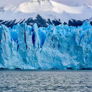A massive blue glacier rises from the dark water, with snow-covered mountains in the background.