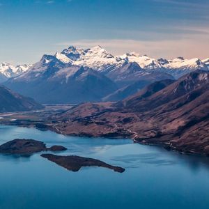 Vue aérienne d'un grand lac bleu avec de petites îles, entouré de montagnes escarpées et de sommets enneigés en arrière-plan.