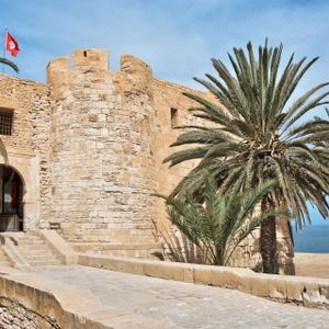 Eine sandfarbene Steinfestung mit einer tunesischen Flagge steht neben einem Strand mit Palmen und dem Meer im Hintergrund.