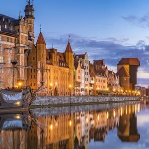 Un galion d'époque est amarré le long d'un canal de la ville au crépuscule, avec des bâtiments historiques illuminés se reflétant dans l'eau calme.