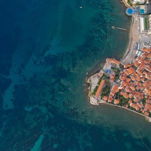 Una veduta aerea, dall'alto, di una città costiera con tetti in tegole arancioni che si affaccia su acque turchesi e limpide, accanto a un porto turistico.