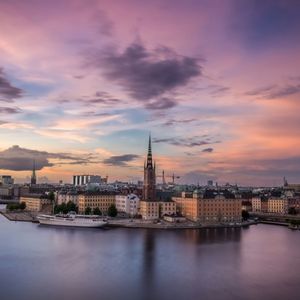 Skyline di una città europea sul lungomare con un campanile prominente, sotto un vibrante tramonto rosa e viola.