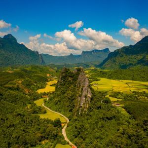 Une vue aérienne d'une route sinueuse à travers une vallée verdoyante avec des rizières, entourée de majestueuses montagnes calcaires sous un ciel bleu.