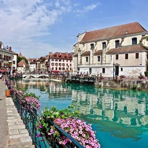 A turquoise canal flows through a historic town, with people strolling on the riverside promenade and buildings reflected in the water.