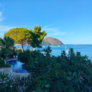 Una piscina immersa nel verde lussureggiante si affaccia su un mare blu con barche e un'isola lontana sotto un cielo sereno.