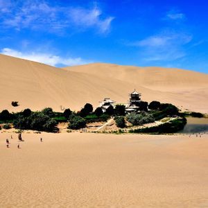 Un'oasi con un lago a mezzaluna e una pagoda tradizionale è incastonata tra vaste dune di sabbia del deserto sotto un cielo azzurro brillante.