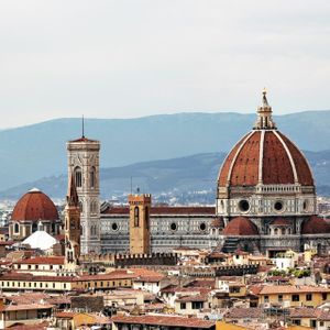 Un paesaggio urbano con tetti in terracotta dominato da una grande cattedrale con una cupola di tegole rosse e un alto campanile, con montagne sullo sfondo.