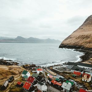 Vue plongeante d'un petit village côtier aux maisons colorées, niché entre une grande montagne herbeuse et la mer, sous un ciel nuageux.
