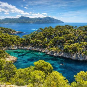 Una vista dall'alto di una caletta rocciosa con acqua cristallina turchese, circondata da rigogliosi pini verdi, con montagne oltre il mare.