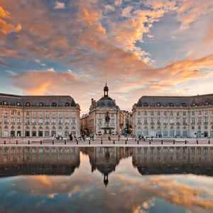 Un grand bâtiment historique se reflète parfaitement dans un miroir d'eau sur une place de ville sous un ciel de coucher de soleil coloré.