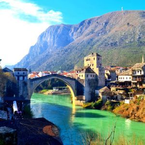 Eine historische Steinbogenbrücke überquert einen türkisfarbenen Fluss, der durch eine Altstadt am Fuße eines großen Berges unter blauem Himmel fließt.