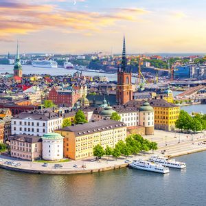 Una vista aerea di una storica città europea con edifici colorati e un campanile, situata su un lungomare con barche ormeggiate al tramonto.