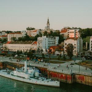 Vista aérea de un buque de guerra blanco atracado en un paseo marítimo en una ciudad, con un campanario de iglesia en una colina al fondo.
