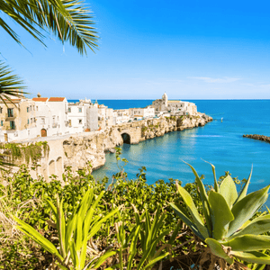 Una località costiera con edifici bianchi si adagia su scogliere rocciose a picco su un mare blu e calmo, con la vista incorniciata da verdeggiante fogliame e foglie di palma.
