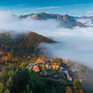 Vue aérienne d'un petit village à flanc de montagne, couvert d'arbres aux couleurs automnales, émergeant d'une mer de nuages.