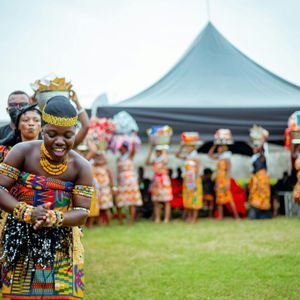 Una donna con abiti tradizionali colorati e gioielli di perline sorride mentre balla su un prato, guidando un corteo di persone.