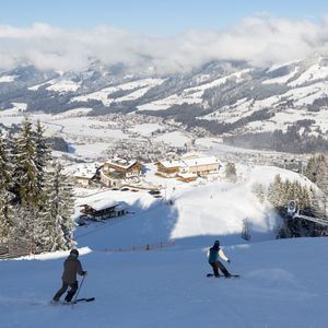 A skier and a snowboarder from a WeRoad group trip descend a sunny slope above a snow-covered mountain village.