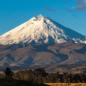 Un gran volcán cubierto de nieve emite una columna de humo desde su cima, dominando un paisaje de colinas y árboles bajo un cielo azul claro.