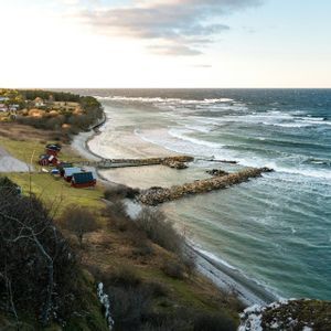Blick von oben auf ein Küstendorf mit roten Häusern an einem grasbewachsenen Ufer, neben einem unruhigen Meer mit weißen Wellen.