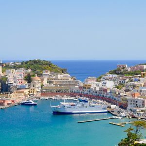 Case colorate su una collina circondano un porto blu brillante dove un grande traghetto e barche più piccole sono ormeggiate sotto un cielo sereno.