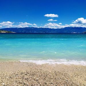 Una spiaggia di ciottoli si affaccia su acque turchesi vibranti, con montagne in lontananza sotto un cielo azzurro brillante e nuvole sparse.