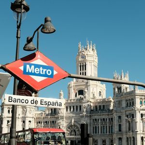 A red Metro sign for the Banco de España station, with a red tour bus and an ornate white building under a clear blue sky.