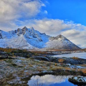 Un paesaggio di montagne innevate che si ergono sopra un villaggio costiero e un fiordo, con un primo piano roccioso, parzialmente innevato.