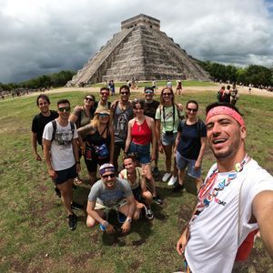 Un gruppo WeRoad si scatta un selfie sorridente su un campo erboso di fronte a una grande antica piramide di pietra sotto un cielo nuvoloso.