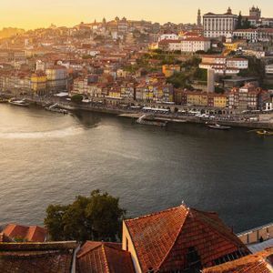 Ein Panoramablick auf eine Stadt am Fluss bei Sonnenuntergang, mit einer großen Bogenbrücke und bunten Häusern am Hang.