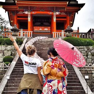 A WeRoad group leader stands with a woman in a traditional kimono and parasol at the base of stairs leading to an orange temple.