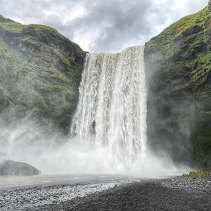 A powerful waterfall cascades down a green, mossy cliff onto a dark pebble shore, creating a thick mist under a cloudy sky.