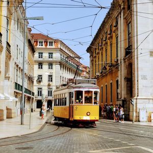 Eine gelbe Straßenbahn fährt auf Gleisen entlang einer Kopfsteinpflasterstraße, umrahmt von historischen Gebäuden mit Bögen unter einem klaren blauen Himmel.