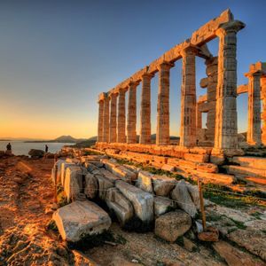 Antiche rovine di templi con grandi colonne di pietra su una scogliera rocciosa, illuminate dal sole al tramonto sul mare.