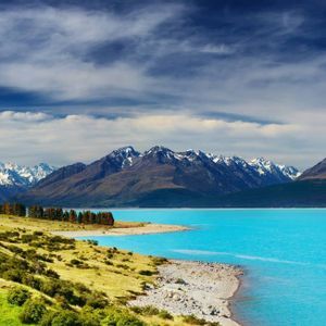 Un vivace lago turchese incontra una sponda erbosa, con una catena di montagne innevate sullo sfondo sotto un cielo nuvoloso.