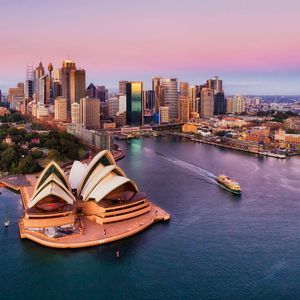 An aerial view of an iconic opera house and a city skyline at sunset, with a ferry crossing the harbor.
