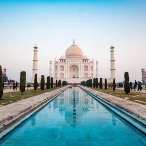 Un largo espejo de agua conduce a un gran mausoleo de mármol blanco con minaretes, rodeado de jardines donde las personas pasean.