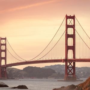 Eine große rote Hängebrücke überspannt ein Gewässer mit Hügeln im Hintergrund unter einem warm getönten Himmel bei Sonnenuntergang.