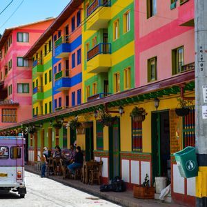 Une rue pavée bordée de bâtiments rayés aux couleurs vives, où un véhicule violet est stationné et des personnes sont attablées en terrasse.