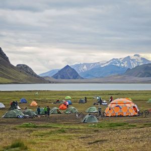 Ein großer Campingplatz mit farbenfrohen Zelten und Fahrrädern auf einer grasbewachsenen Ebene neben einem See, mit schroffen Bergen im Hintergrund unter einem bewölkten Himmel.