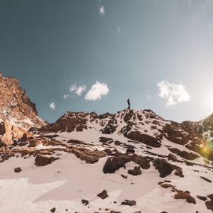 A lone person stands on the summit of a rocky, snow-covered mountain under a bright sun and blue sky.