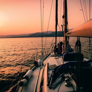 Eine WeRoad-Gruppenreise an Deck eines Segelboots, die einen orangefarbenen Sonnenuntergang über dem Wasser mit fernen Bergen genießt.