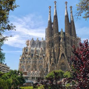 A large, ornate cathedral with tall spires under construction, seen through green and red foliage against a blue sky.