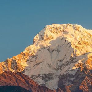 Una grande cima montuosa coperta di neve illuminata dalla luce dorata del sole contro un cielo azzurro e limpido.