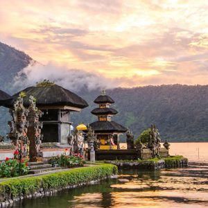 Un complesso templare a più livelli si trova sulla riva di un lago calmo, con montagne boscose sullo sfondo sotto un cielo al tramonto.